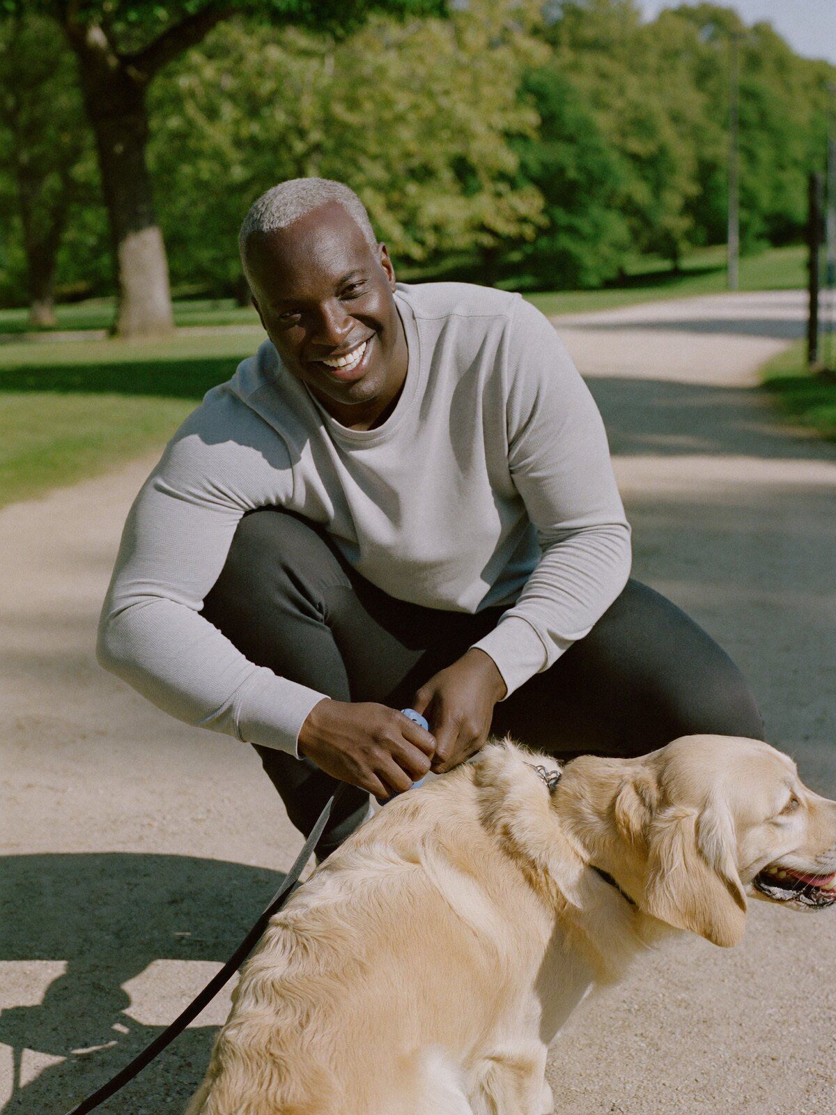 A man crouching on a park path to clip a lead onto a golden retriever